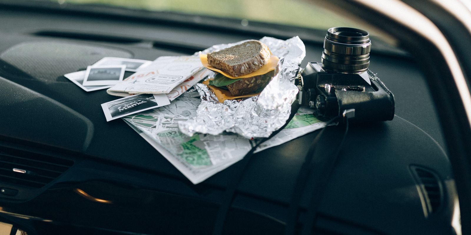 A camera, a foil-wrapped sandwich, folded paper maps and photos on a car dashboard