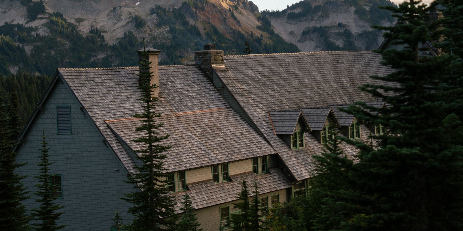 Steep shingled roof of a historic lodge among fir trees with mountain slopes behind at dusk
