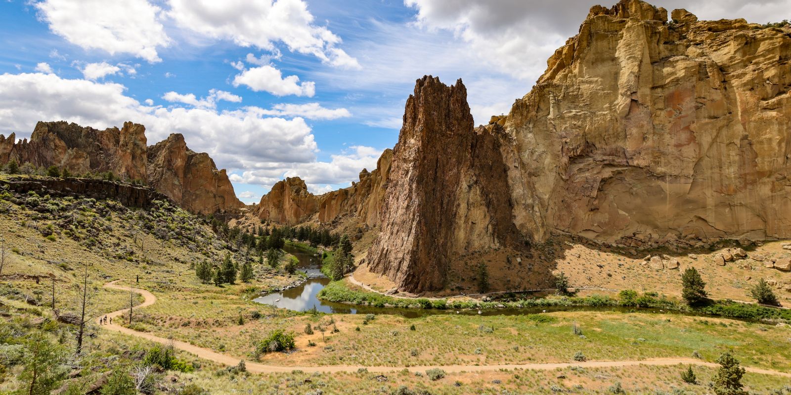 Tall tan rock spires of Smith Rock State Park above a bend in the river and a dirt trail through dry grass.