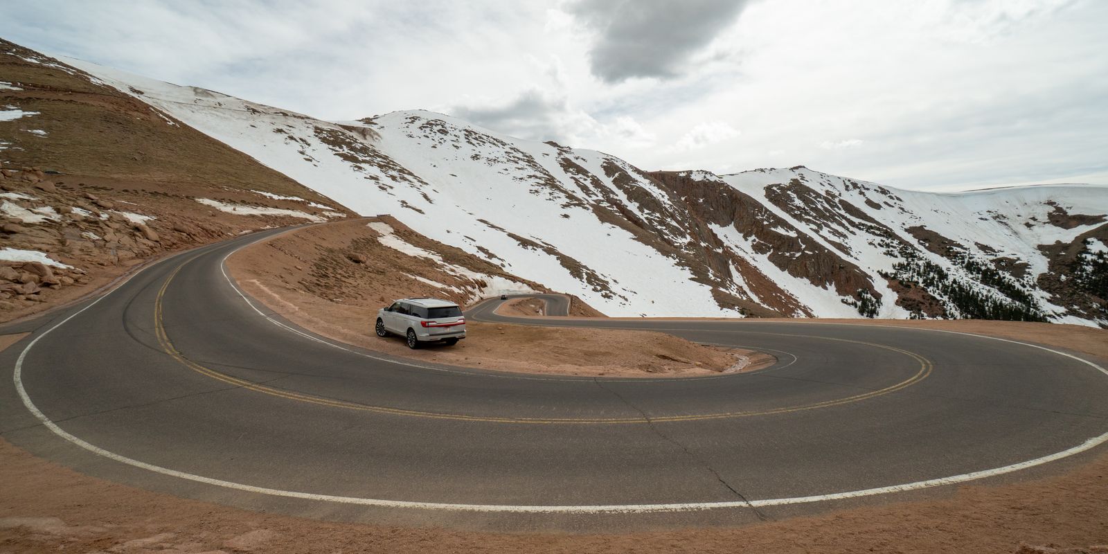 A paved switchback on the Pikes Peak Highway curving below snowy slopes, a single white car on the bend.
