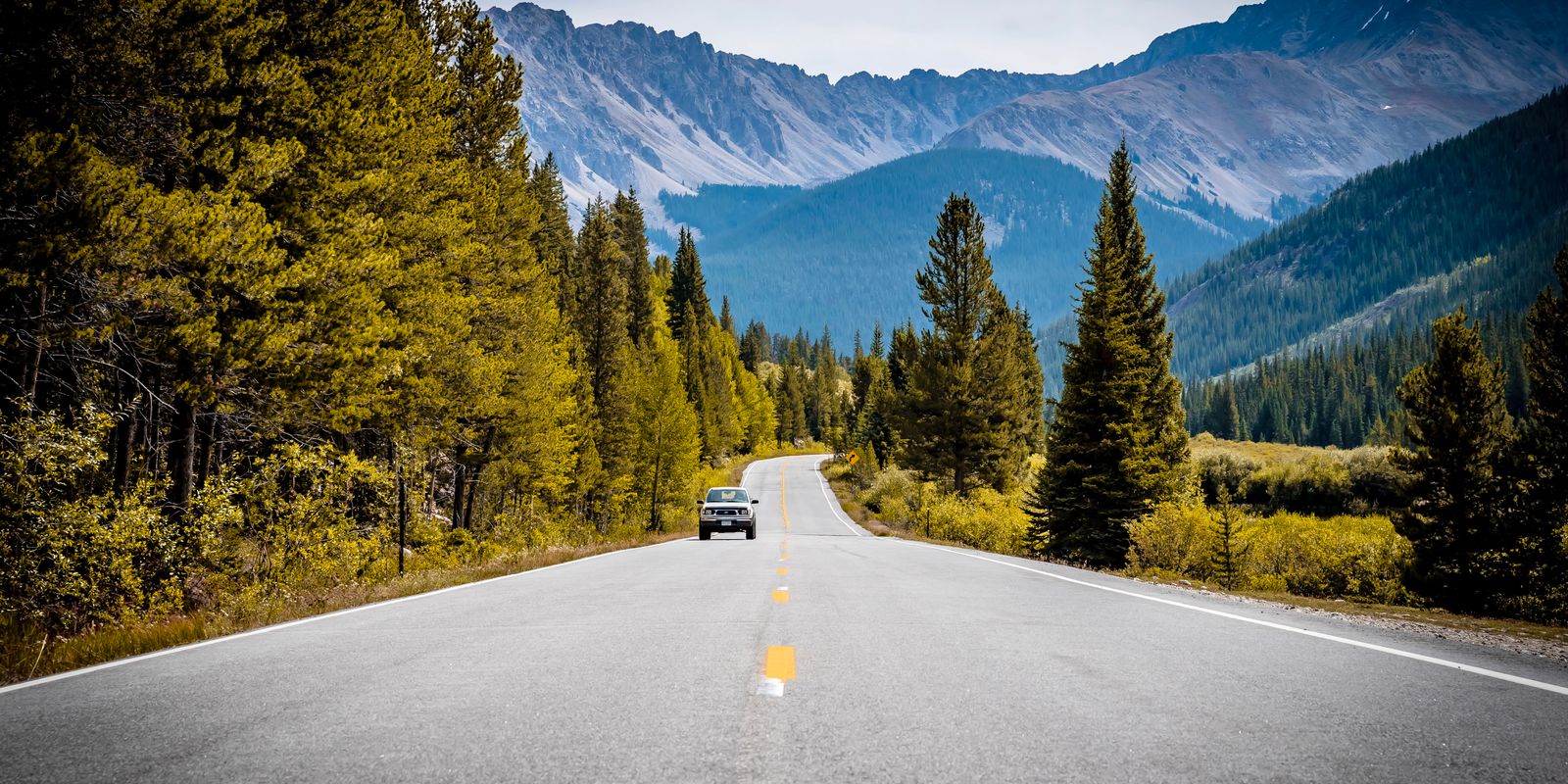 Two-lane road through pine and yellowing aspen toward rocky Colorado peaks, one car ahead.