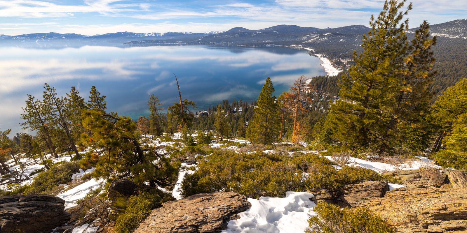 Lake Tahoe seen through pines from a rocky slope with patches of snow