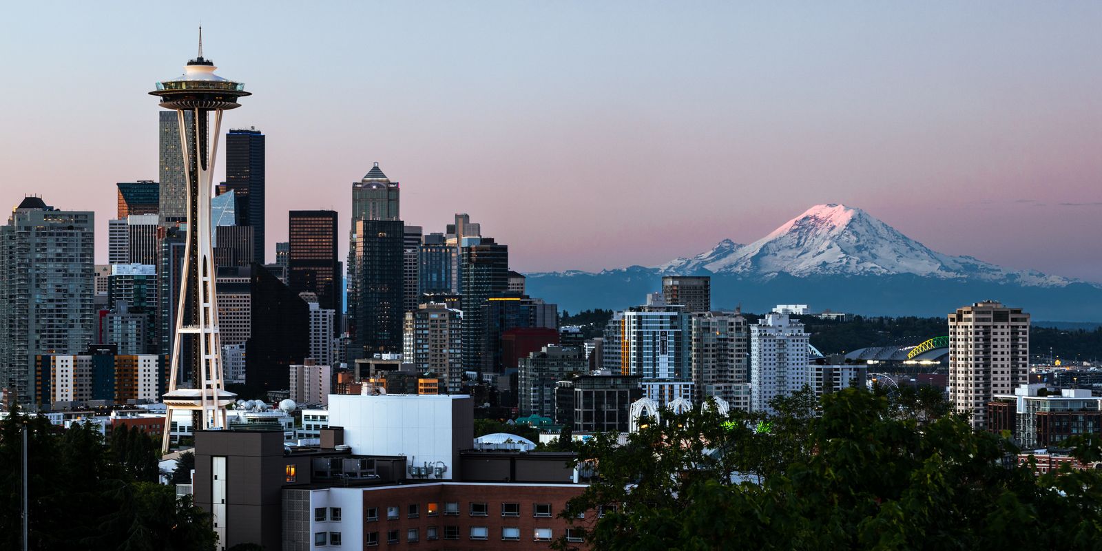 Seattle skyline at dawn with the Space Needle on the left and snow-covered Mount Rainier glowing pink in the distance.