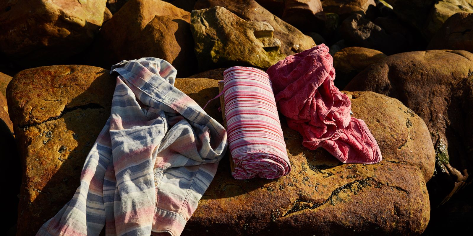 A striped shirt, a rolled towel and a red towel lying on sunlit boulders