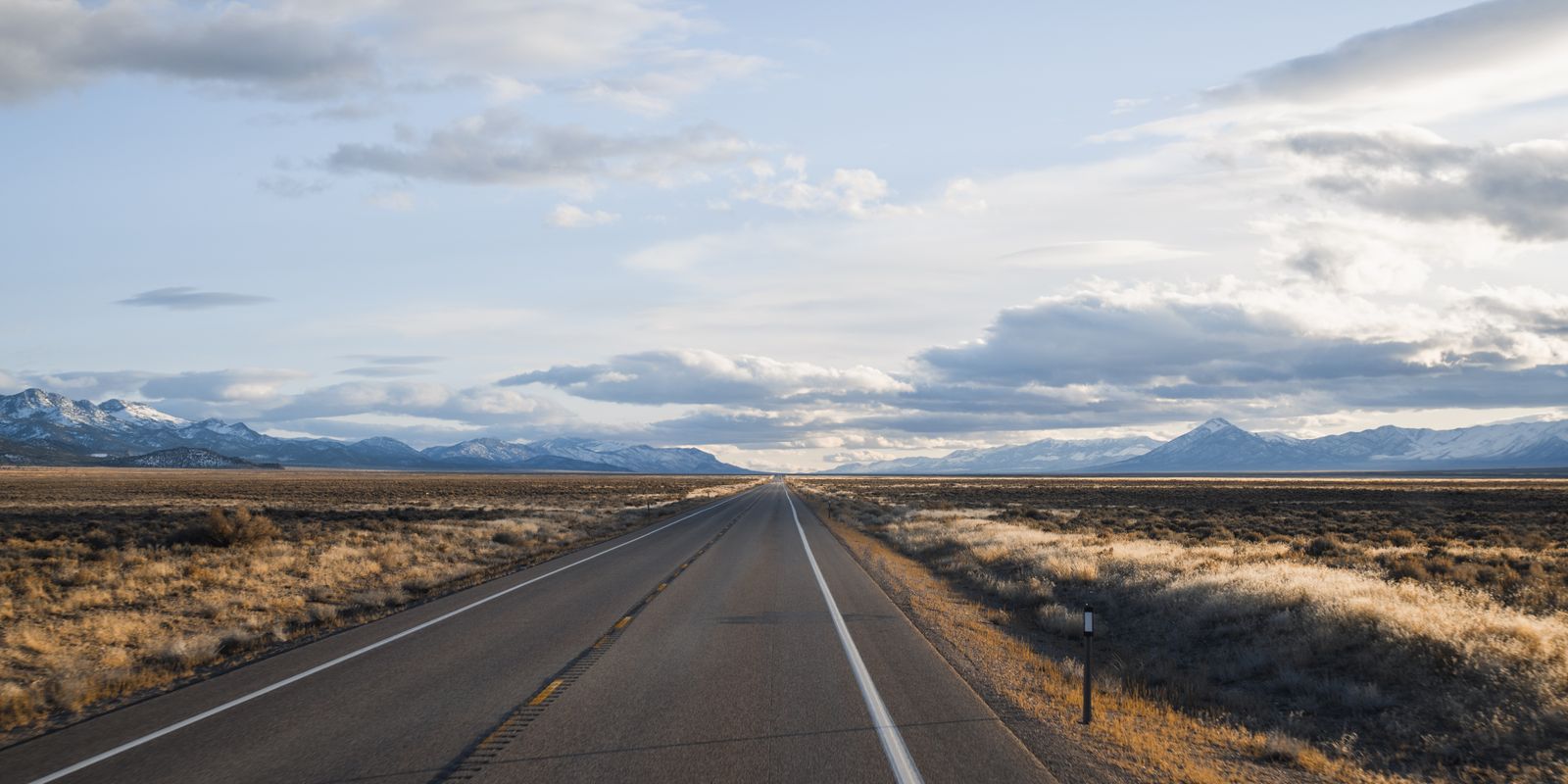 Empty two-lane highway across Nevada grassland toward snow-capped mountains under a cloudy sky