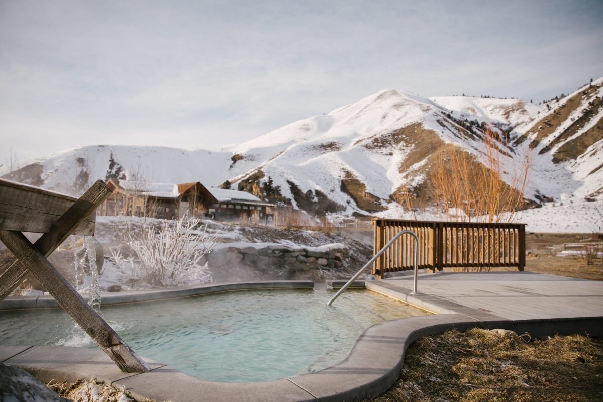 Steaming soaking pool with a wooden spout, snow-covered hills and lodge buildings behind
