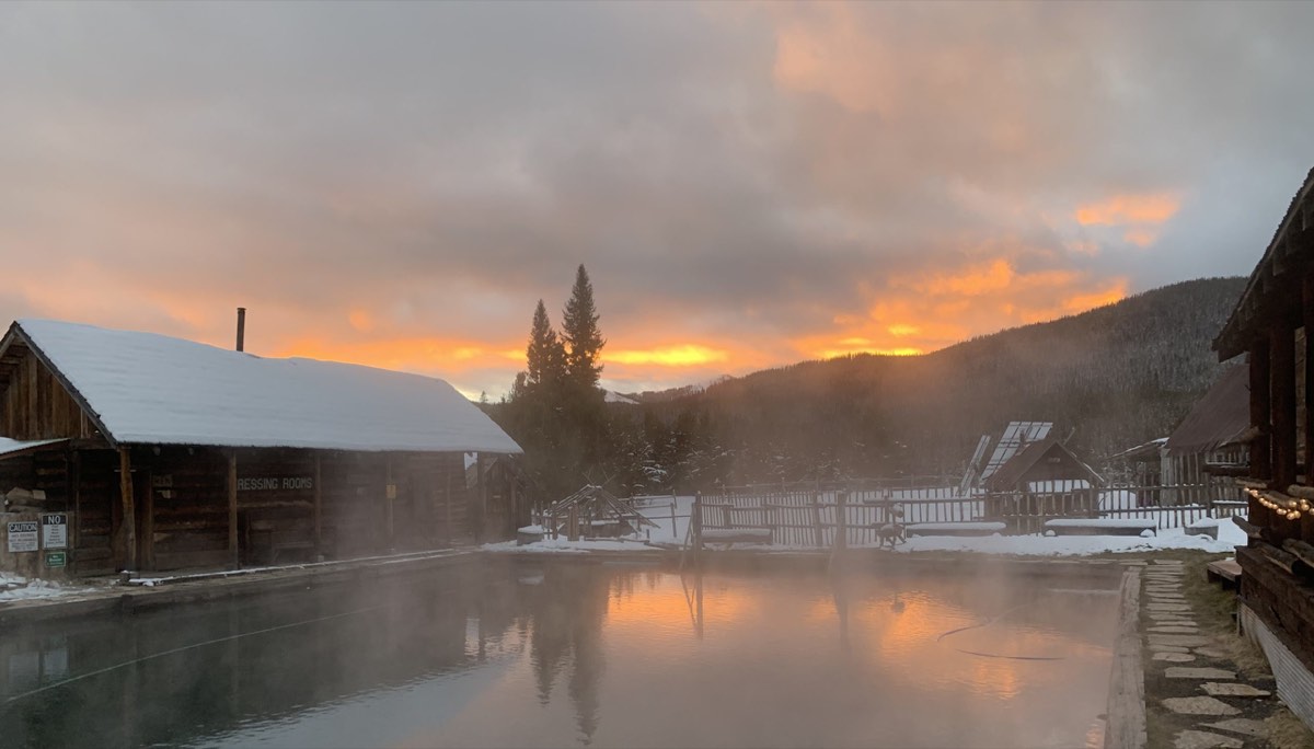 Steaming log-edged pool beside a snowy dressing-room cabin at sunset, forested hills behind