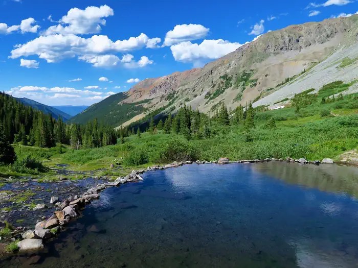Conundrum Hot Springs' stone-rimmed pool in a green alpine meadow below rocky peaks