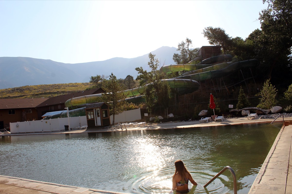 Large outdoor mineral pool with green waterslides and mountains behind, one soaker seen from behind