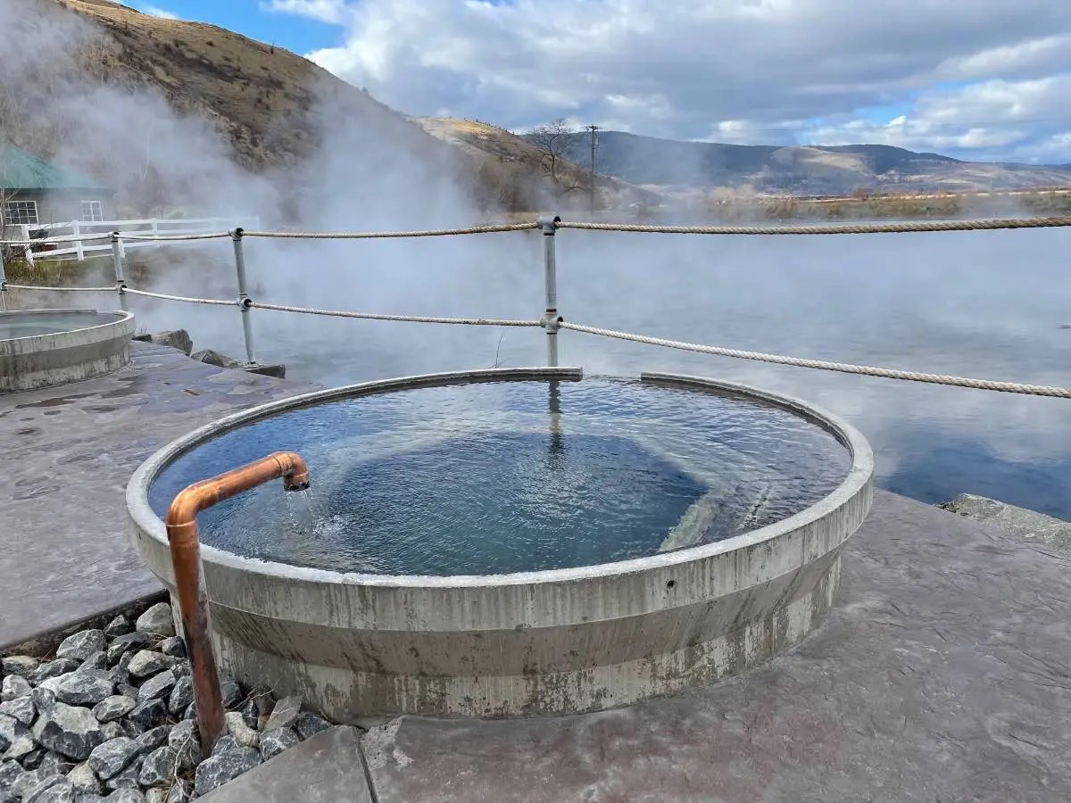 Steaming round concrete soaking tub with a copper spout on the lakeshore, hills behind
