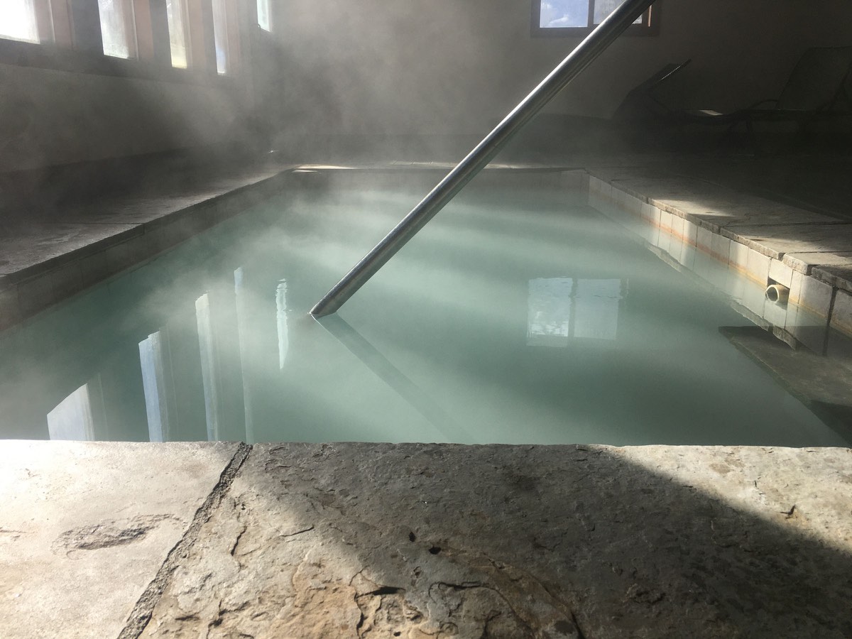 Steaming milky-blue mineral pool inside a stone-floored bathhouse with tall windows