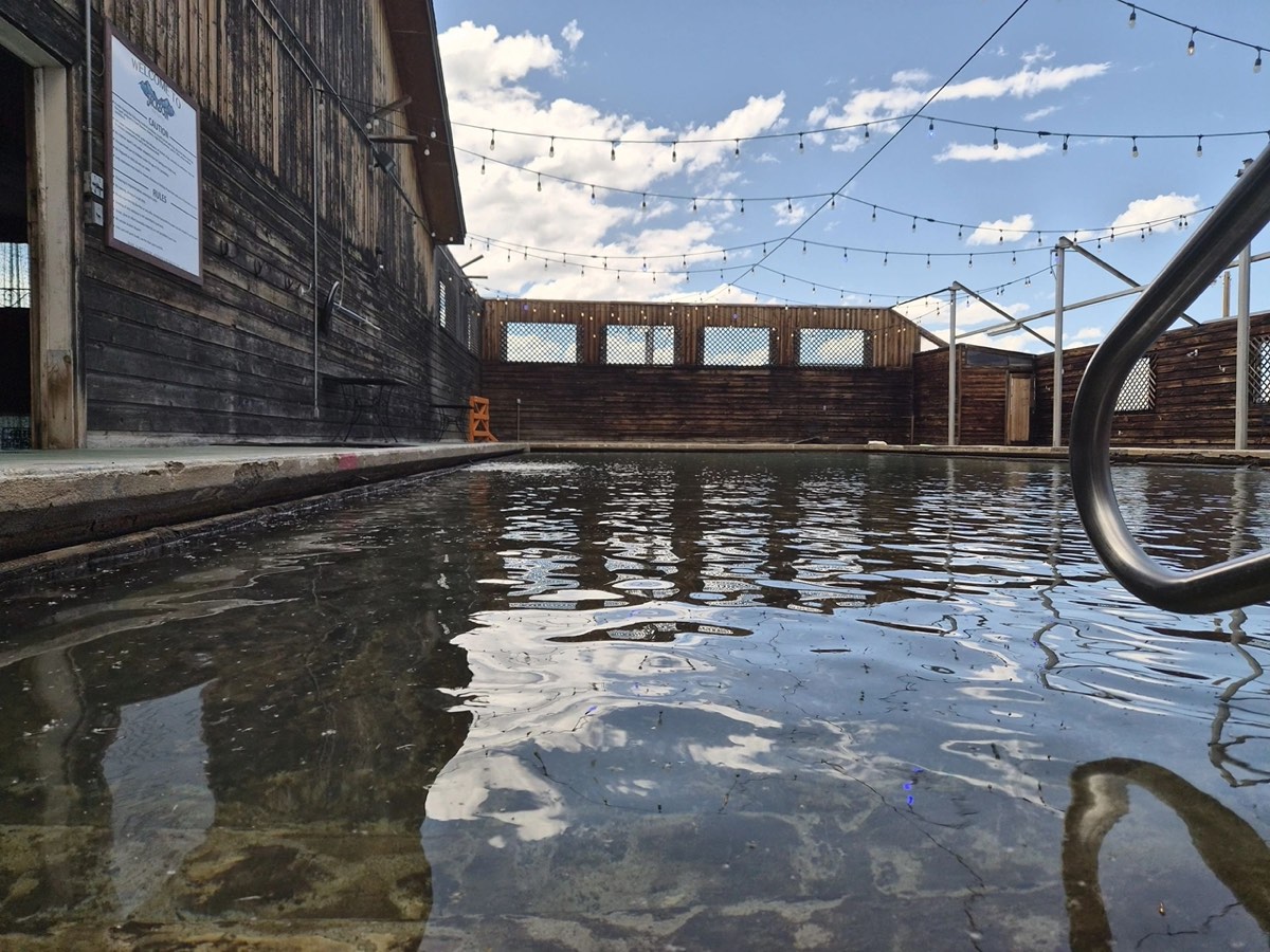 Water-level view of the outdoor hot spring pool beside the weathered wood lodge under string lights