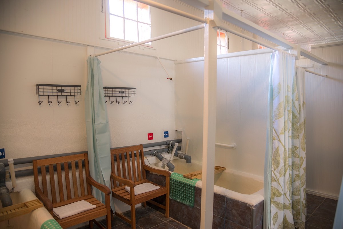 Indoor tiled soaking tubs with hot and cold taps, wooden chairs and shower curtains