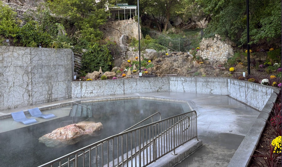 Steaming outdoor hot pool with a boulder island and handrails below a rocky, flowered hillside