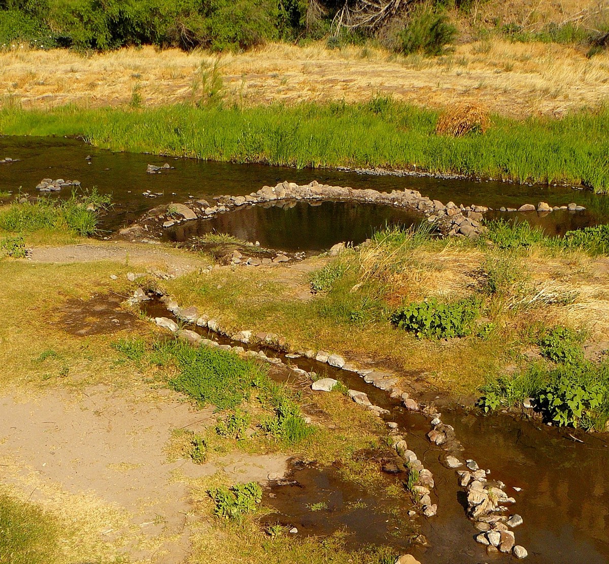 Rock-ringed hot spring pool and stone-lined runoff channel in a grassy meadow beside the Gila Middle Fork