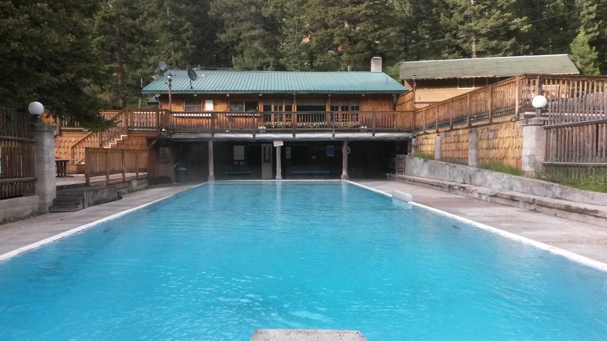 Long blue spring-fed outdoor pool in front of a wooden lodge with decks, pines behind
