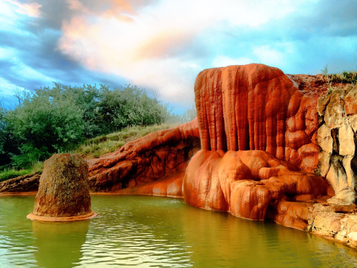 Orange travertine mound spilling into a green spring-fed pool under a cloudy sky