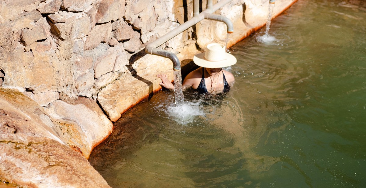 A soaker in a sun hat under a pipe spout in Ojo Caliente's stone-walled iron pool