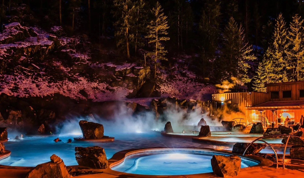 Steaming lit pools among boulders at night beside a timber bathhouse in a snowy canyon