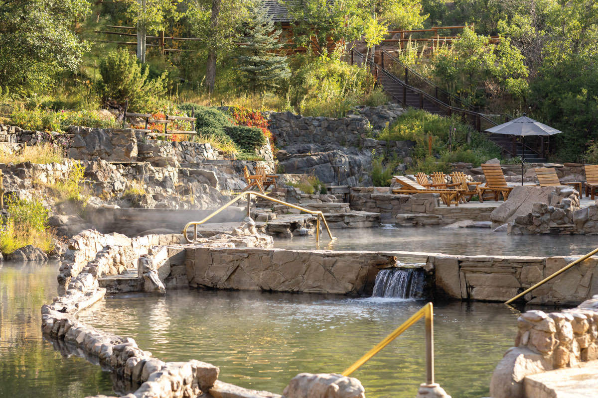 Stone masonry hot pools with a small waterfall, steam and lounge chairs on a wooded hillside